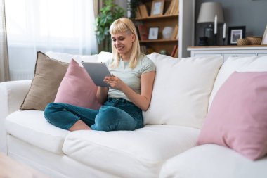 Evdeki kanepede dijital tablet teknolojisi kullanan mutlu genç kız bilgisayarını tutuyor. Uygulamaları, alışverişi, online randevuları, haberleri, kanepede interneti kullanarak gülümseyen genç kadın..