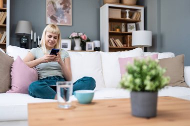 Mutlu, rahat, genç bir kadın koltukta oturmuş telefonla gülüyor, elinde akıllı telefonuyla gülüyor, cep telefonuna bakıyor mobil uygulamalarla online ekommerce alışverişi yapıyor ya da internetten flört ediyor..
