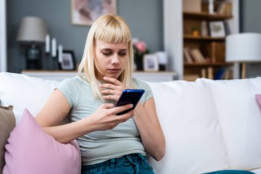 Mutlu, rahat, genç bir kadın koltukta oturmuş telefonla gülüyor, elinde akıllı telefonuyla gülüyor, cep telefonuna bakıyor mobil uygulamalarla online ekommerce alışverişi yapıyor ya da internetten flört ediyor..