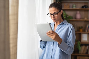 İş dünyasındaki zeki beyaz kadın evdeki pencerelerin yanında elinde dijital tablet tutuyordu. Genç bağımsız kadın işçi teknolojiyi ev işlerinde uzak işlerde kullanıyor..