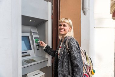 Young backpacker girl withdraws cash money from an ATM machine with her credit card to continue traveling and visiting destinations and countries and cities around the world with her free lifestyle