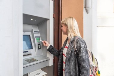 Genç bir sırt çantalı kız ATM 'den nakit para çekiyor. Kredi kartıyla seyahat ve tatil beldelerini, ülkeleri ve dünyadaki şehirleri bedava yaşam tarzıyla ziyaret ediyor.