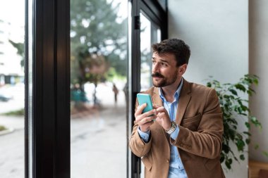 Ofiste dikilip Büyük Şehir 'de pencereden dışarı bakan akıllı telefonu kullanan düşünceli bir iş adamı. E-Ticaret Girişimi için Karizmatik Dijital Girişimci Planlama Yatırım Stratejisi. İş Fikirleri