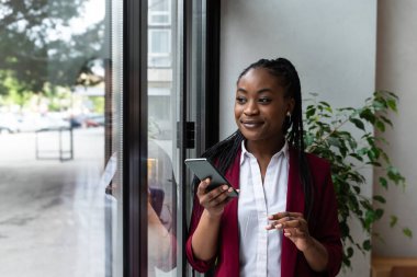 Akıllı telefon kullanan düşünceli iş kadını ofiste dikilip Büyük Şehir 'de pencereden dışarı bakıyor. E-Ticaret Girişimi için Karizmatik Dijital Girişimci Planlama Yatırım Stratejisi. İş Fikirleri
