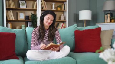 Evdeki kanepede dinlenen genç bir kadın. Kablosuz kulaklıklardan müzik dinlemek ve kitap okumak. Kitap seven kadın, rahat dairesinde dinlenmek için boş zaman ve hafta sonu aktivitelerinin tadını çıkarıyor.