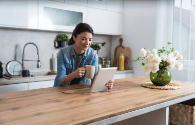 Gülümseyen, gözlüklü, kahve içen ve mutfakta otururken tablet bilgisayar kullanan genç bir kadın. İş kadını sabahları evinde e-posta okuyor. Uzak iş kadınları