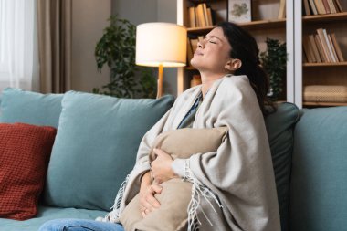 Regl olmuş genç bir kadın kanepeye uzanıyor ve karnına tutunuyor çünkü regl döneminin ortasında acı dolu bir yüz ifadesi ile acı çekiyor.