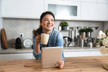 Gülümseyen başarılı genç kadın mutfakta otururken kahve içip tablet bilgisayar kullanıyor. İş kadını sabahları evinde e-posta okuyor. Uzak iş sahibi kadın.