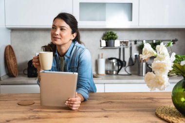 Gülümseyen başarılı genç kadın mutfakta otururken kahve içip tablet bilgisayar kullanıyor. İş kadını sabahları evinde e-posta okuyor. Uzak iş sahibi kadın.