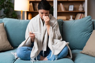 Hastalık, mevsimsel virüs problemi konsepti. Kanepede yatıp termometredeki sıcaklığa bakarak hasta olmak. Hasta kadın yüksek ateşle battaniyeye sarılı yatıyordu. Soğuk algınlığı ve migren.