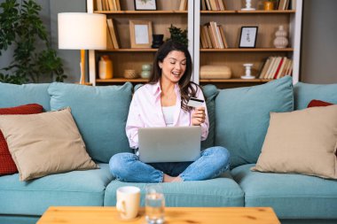 Genç, mutlu, bağımsız bir kadın kanepeye oturmuş alışveriş yapıyor. Banka kartlı dizüstü bilgisayardan online alışveriş..