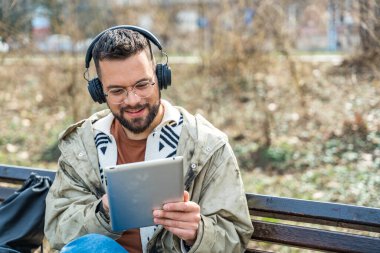 Havalı tavırlı genç hippi işadamı parkta oturmuş dijital tabletle video görüşmesi yapıyor. Kalabalığın dışında duran gurbetçi iş adamı, akıllı tasarım profesyonel mesleği.