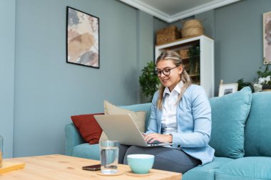 Genç ve güzel bir iş kadını evinde dizüstü bilgisayarını kullanarak kanepede uzanıyor. İnternette çalışıyor, video görüşmesi yapıyor. Yaratıcı kadın gülümsemesi, sosyal medyayı kontrol etme, mesaj yazma.