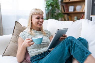 Dijital tablet kullanan çekici kadın serbest yazar oturma odasındaki rahat koltukta oturuyor. Başarılı bir iş kadını evde mesaj atıyor. Çevrimiçi teknoloji konsepti, uzak iş e-ticaret e-öğrenimi.