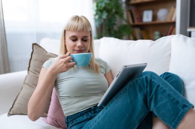 Dijital tablet kullanan çekici kadın serbest yazar oturma odasındaki rahat koltukta oturuyor. Başarılı bir iş kadını evde mesaj atıyor. Çevrimiçi teknoloji konsepti, uzak iş e-ticaret e-öğrenimi.