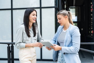 Modern ofis binasının dışında dijital tablet kullanan iki iş kadını yeni proje üzerinde çalışıyor iş konularını tartışıyor ve açıklıyor ve gülümsüyor. Profesyonel işgalci kişiler tasarla