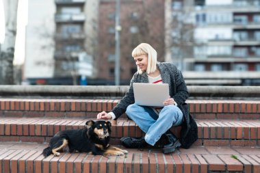 Profesyonel tasarım. Serbest çalışan genç bir iş kadını, bilgisayar yazılımı geliştiricisi, web tasarımcısı, evcil köpeği ve dizüstü bilgisayarıyla dışarıda otururken uzaktan çalışıyor..