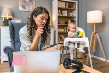 Genç bir kadın, anne ve iş kadını, dizüstü bilgisayarında kredi kartıyla evde alışveriş yaparken, bebeği bebekler için bebek koltuğunda oturup oyuncaklarla oynuyor. Anne ve çocuk evde..
