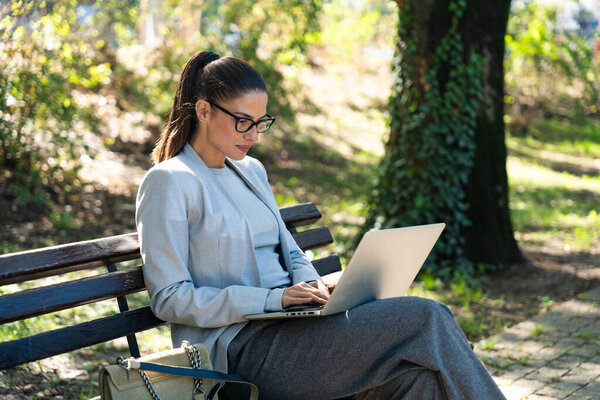 Прекрасная улыбчивая бизнесвумен в формальной одежде, сидящая на лавочке под открытым небом в парке и планирующая с помощью ноутбука работать или проводить онлайн-встречи. Бизнес, финансовая индустрия или концепция недвижимости.