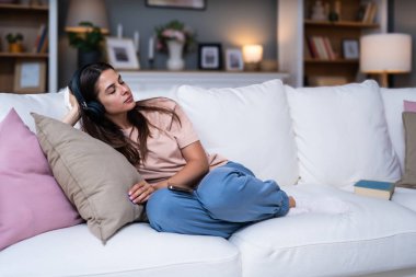 Kendini sevme zamanı. Genç ve güzel bir kadın küçük evinde dinleniyor ve dinleniyor modern stüdyo dairesinde, kablosuz kulaklıklarıyla müzik dinliyor, kanepede otururken. Hafta sonu aktiviteleri.