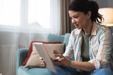Çekici kadın, serbest çalışan, dijital tablet kullanan, oturma odasındaki rahat koltukta oturan. Başarılı bir iş kadını evde mesaj atıyor. Çevrimiçi teknoloji konsepti, uzak meslek.