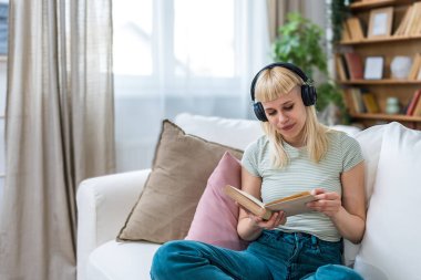 Gündelik kıyafetlerle kanepede uzanan, kablosuz kulaklıklarla müzik dinleyen, kitap okuyan genç bir kadın. Okuma zamanı, kitap seven kadın hafta sonu aktivitelerinin keyfini çıkarıyor..