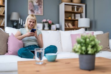 Gülümseyen bir kadın akıllı telefon ekranına bakıyor, koltukta oturuyor, evdeki mobil cihazlarla eğleniyor, sosyal ağda sohbet ediyor, internette sörf yapıyor, internetten alışveriş yapıyor.