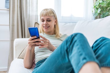 Gülümseyen bir kadın akıllı telefon ekranına bakıyor, koltukta oturuyor, evdeki mobil cihazlarla eğleniyor, sosyal ağda sohbet ediyor, internette sörf yapıyor, internetten alışveriş yapıyor.