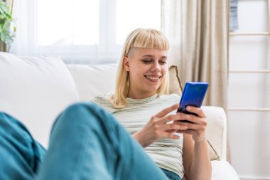 Gülümseyen bir kadın akıllı telefon ekranına bakıyor, koltukta oturuyor, evdeki mobil cihazlarla eğleniyor, sosyal ağda sohbet ediyor, internette sörf yapıyor, internetten alışveriş yapıyor.