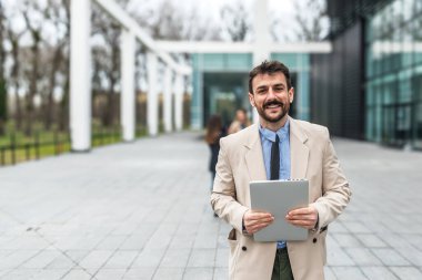 Ofis binasının önünde dijital tablet kullanarak şık kıyafetler giyen başarılı genç işadamının portresi. Profesyonel mesleki erkek, ofisten personel toplantısına yürürken çalışıyor..