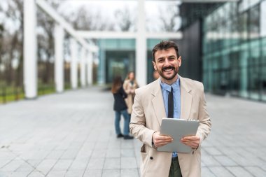 Ofis binasının önünde dijital tablet kullanarak şık kıyafetler giyen başarılı genç işadamının portresi. Profesyonel mesleki erkek, ofisten personel toplantısına yürürken çalışıyor..