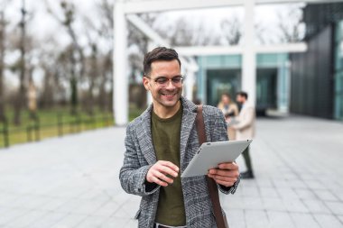 Ofis binasının önünde dijital tablet kullanarak şık kıyafetler giyen başarılı genç işadamının portresi. Profesyonel mesleki erkek, ofisten personel toplantısına yürürken çalışıyor..