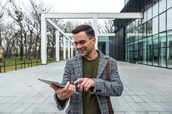 Ofis binasının önünde dijital tablet kullanarak şık kıyafetler giyen başarılı genç işadamının portresi. Profesyonel mesleki erkek, ofisten personel toplantısına yürürken çalışıyor..