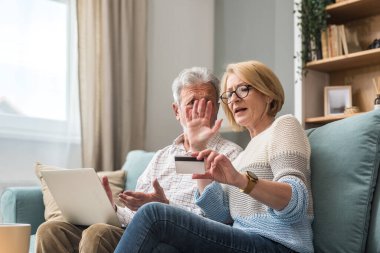 Üst düzey çift, erkek ve kadın, online alışveriş ve sipariş için dizüstü bilgisayar ve banka kredi kartı kullanıyorlar. Evdeki kanepede otururken internet ve teknoloji kullanan evli insanlar..