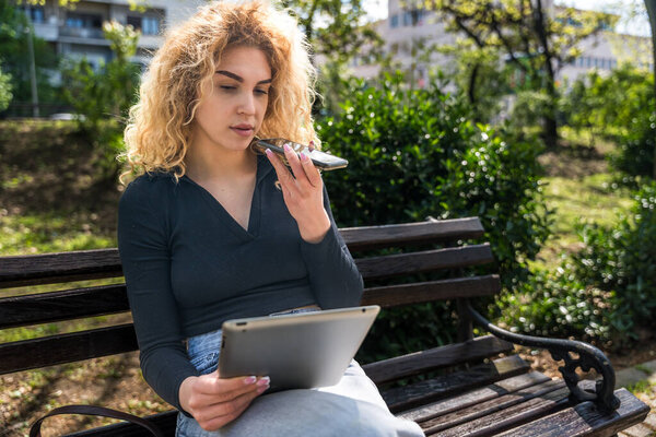 Young university student girl sitting on a park bench, studying and reading material on a digital tablet, preparing for a final exam at college. Checking emails on the device, class schedule.