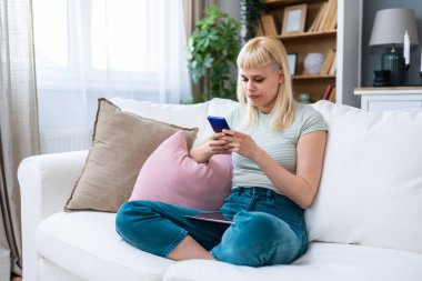 Genç bir kadın rahat koltukta tek başına rahat bir şekilde rahat bir şekilde rahat rahat rahat oturup mesajlaşmak, yayın yapmak, alışveriş yapmak, video sohbeti yapmak, okumak, göz atmak, oyun oynamak, uzaktan çalışmak, bankacılık, öğrenmek, programlamak için akıllı telefon kullanıyor..