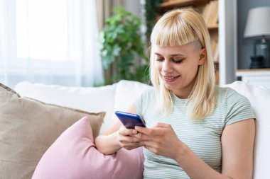 Genç bir kadın rahat koltukta tek başına rahat bir şekilde rahat bir şekilde rahat rahat rahat oturup mesajlaşmak, yayın yapmak, alışveriş yapmak, video sohbeti yapmak, okumak, göz atmak, oyun oynamak, uzaktan çalışmak, bankacılık, öğrenmek, programlamak için akıllı telefon kullanıyor..