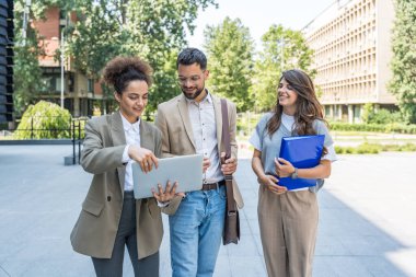 Kurumsal profesyonellerden oluşan çeşitli bir ekip dışarıda iş birliği yapıyor, modern cam ofis binası önünde dizüstü bilgisayardaki verileri analiz ederken stratejik tartışmalara girişiyorlar..