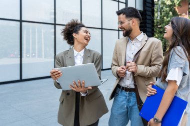 Kurumsal profesyonellerden oluşan çeşitli bir ekip dışarıda iş birliği yapıyor, modern cam ofis binası önünde dizüstü bilgisayardaki verileri analiz ederken stratejik tartışmalara girişiyorlar..