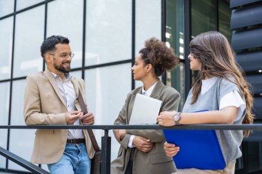 Kurumsal profesyonellerden oluşan çeşitli bir ekip dışarıda iş birliği yapıyor, modern cam ofis binası önünde dizüstü bilgisayardaki verileri analiz ederken stratejik tartışmalara girişiyorlar..