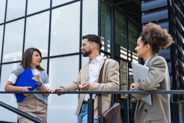 Kurumsal profesyonellerden oluşan çeşitli bir ekip dışarıda iş birliği yapıyor, modern cam ofis binası önünde dizüstü bilgisayardaki verileri analiz ederken stratejik tartışmalara girişiyorlar..