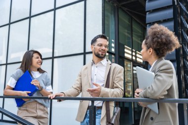 Kurumsal profesyonellerden oluşan çeşitli bir ekip dışarıda iş birliği yapıyor, modern cam ofis binası önünde dizüstü bilgisayardaki verileri analiz ederken stratejik tartışmalara girişiyorlar..