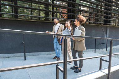 Kurumsal profesyonellerden oluşan çeşitli bir ekip dışarıda iş birliği yapıyor, modern cam ofis binası önünde dizüstü bilgisayardaki verileri analiz ederken stratejik tartışmalara girişiyorlar..