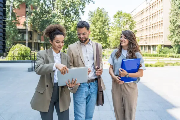 Kurumsal profesyonellerden oluşan çeşitli bir ekip dışarıda iş birliği yapıyor, modern cam ofis binası önünde dizüstü bilgisayardaki verileri analiz ederken stratejik tartışmalara girişiyorlar..