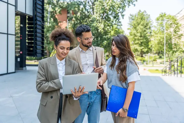Kurumsal profesyonellerden oluşan çeşitli bir ekip dışarıda iş birliği yapıyor, modern cam ofis binası önünde dizüstü bilgisayardaki verileri analiz ederken stratejik tartışmalara girişiyorlar..