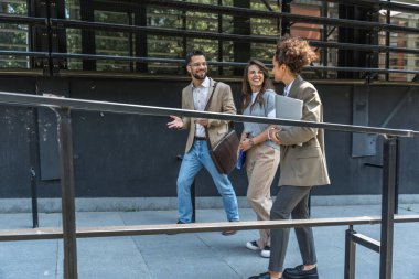 Kurumsal profesyonellerden oluşan çeşitli bir ekip dışarıda iş birliği yapıyor, modern cam ofis binası önünde dizüstü bilgisayardaki verileri analiz ederken stratejik tartışmalara girişiyorlar..