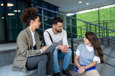 Kurumsal profesyonellerden oluşan çeşitli bir ekip dışarıda iş birliği yapıyor, modern cam ofis binası önünde dizüstü bilgisayardaki verileri analiz ederken stratejik tartışmalara girişiyorlar..