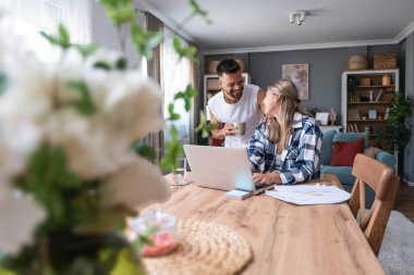 Genç çift evlerinde uzaktan çalışıyor, belge ve verileri analiz ediyor, rahat modern oturma odasında dizüstü bilgisayar kullanıyor, mali raporları ve iş stratejisini tartışıyor. Birkaç hedef, basit yaşam desteği..