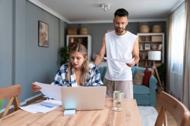 Genç çift evlerinde uzaktan çalışıyor, belge ve verileri analiz ediyor, rahat modern oturma odasında dizüstü bilgisayar kullanıyor, mali raporları ve iş stratejisini tartışıyor. Birkaç hedef, basit yaşam desteği..