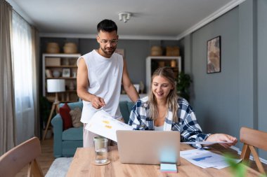 Genç çift evlerinde uzaktan çalışıyor, belge ve verileri analiz ediyor, rahat modern oturma odasında dizüstü bilgisayar kullanıyor, mali raporları ve iş stratejisini tartışıyor. Birkaç hedef, basit yaşam desteği..